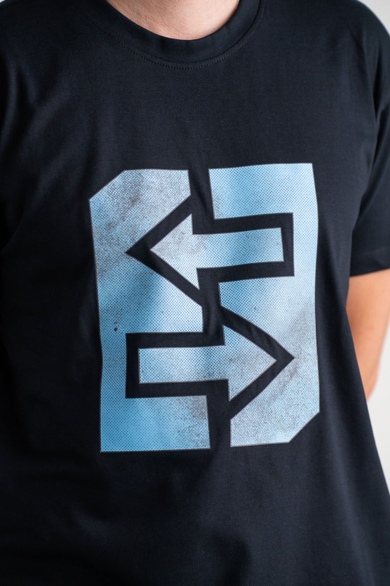 Ein Mann trägt ein schwarzes T-Shirt mit einem blauen Logo, inspiriert von griechischer Mythologie und Geschichte.