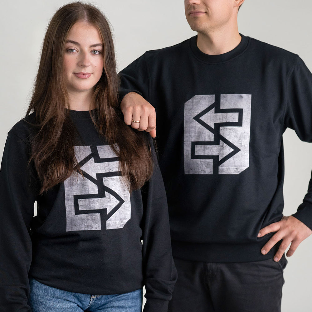 Ein Mann und eine Frau tragen schwarze Sweatshirts mit einem markanten grauen Logo, das Stärke und Unzerstörbarkeit symbolisiert.