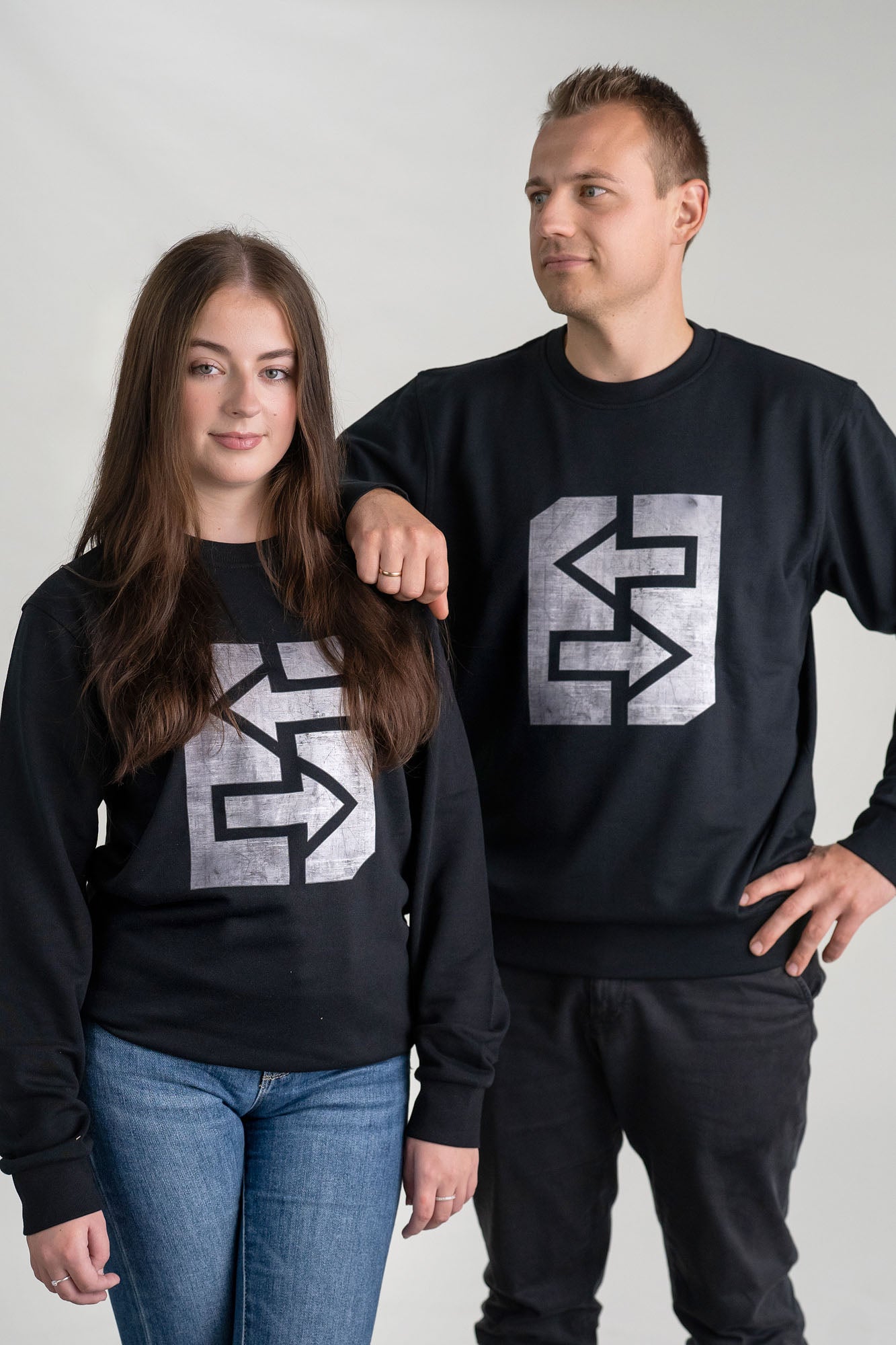 Ein Mann und eine Frau tragen schwarze Sweatshirts mit einem markanten grauen Logo, das Stärke und Unzerstörbarkeit symbolisiert.