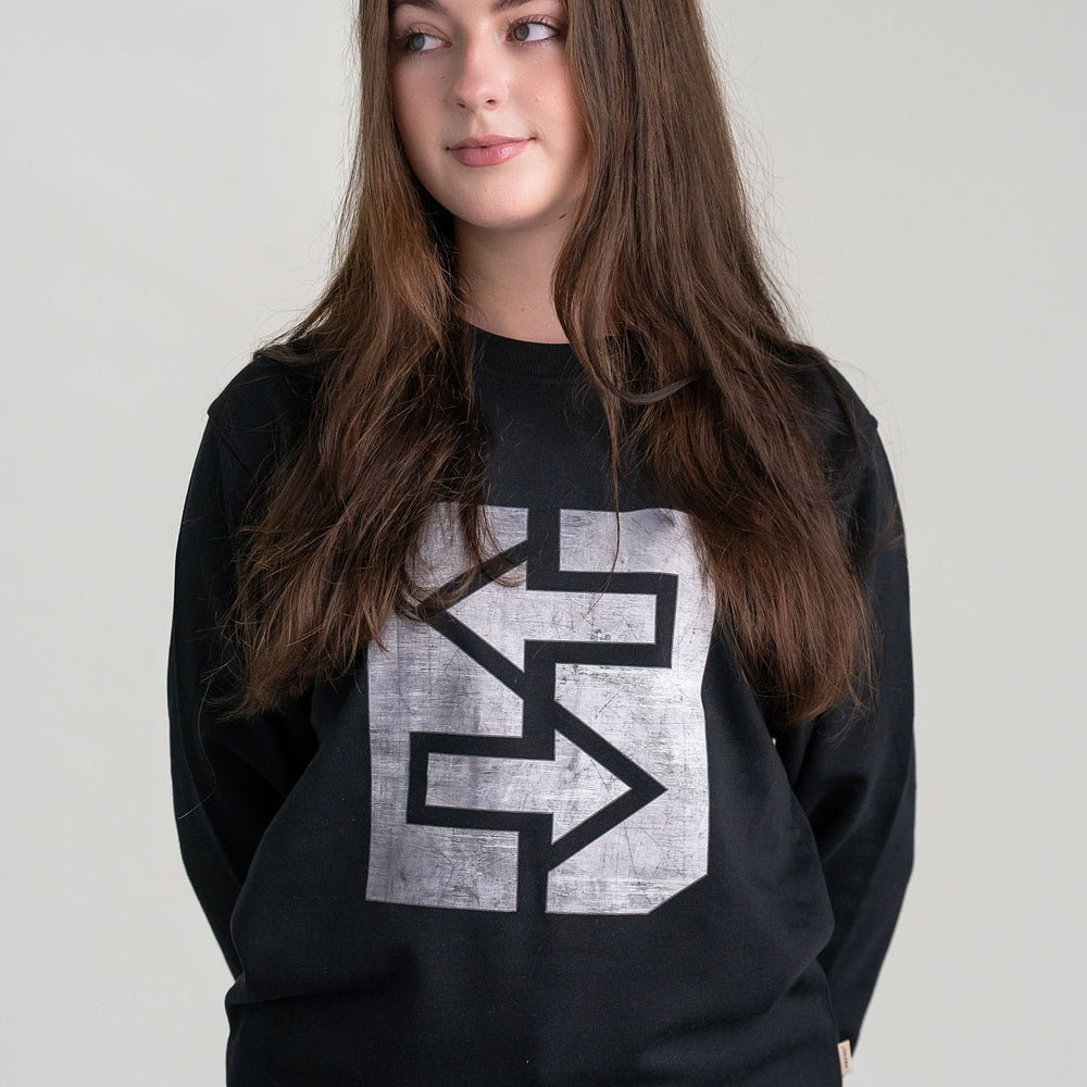 Eine Frau trägt eine schwarze Sweatshirt mit einem markanten grauen Logo, das Stärke und Unzerstörbarkeit symbolisiert.