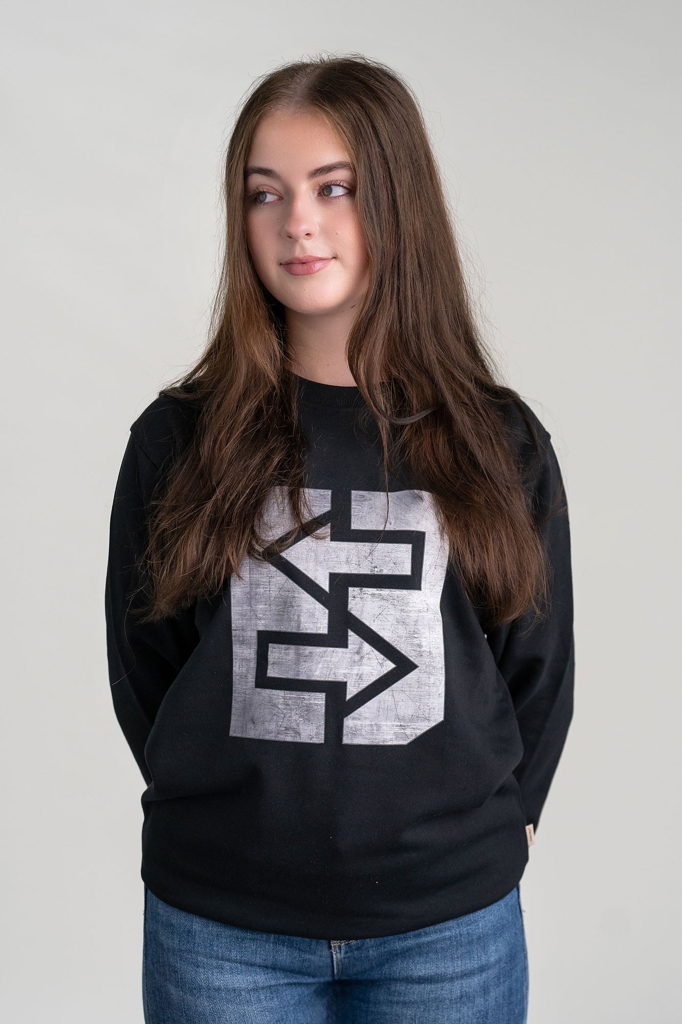 Eine Frau trägt eine schwarze Sweatshirt mit einem markanten grauen Logo, das Stärke und Unzerstörbarkeit symbolisiert.