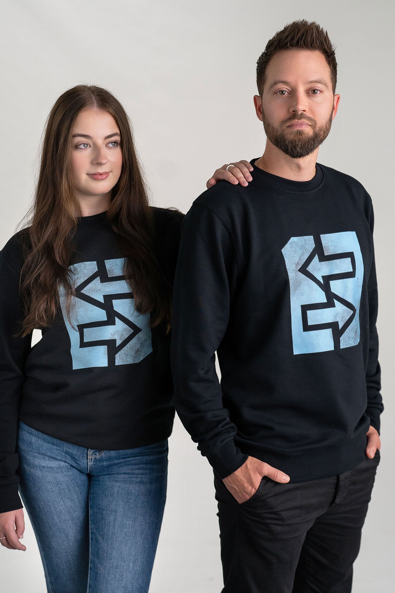 Ein Mann und eine Frau tragen schwarze Sweatshirts mit einem Logo, inspiriert von griechischer Mythologie und Geschichte.