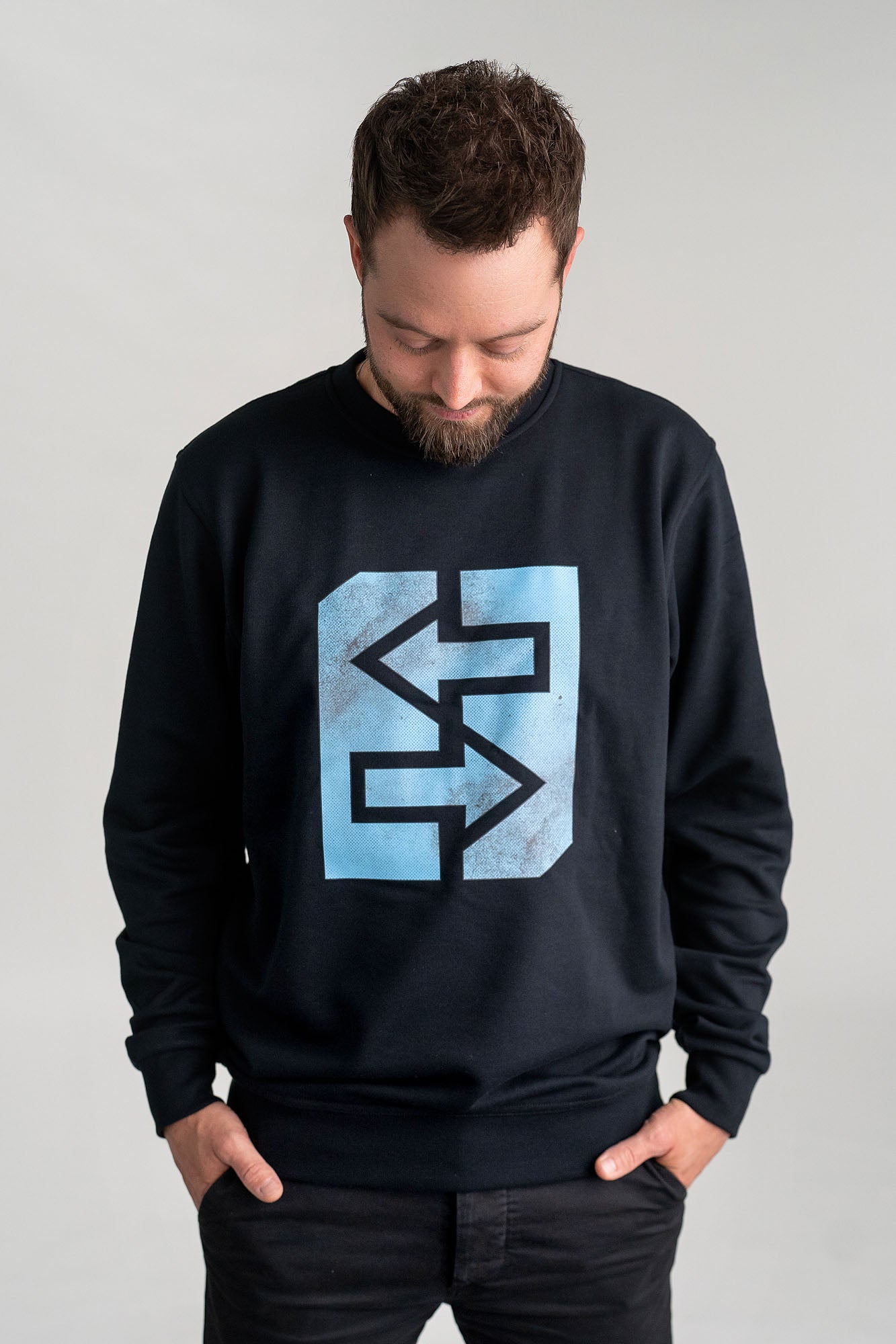 Ein Mann trägt ein schwarzes Sweatshirt mit einem blauen Logo, inspiriert von griechischer Mythologie und Geschichte.
