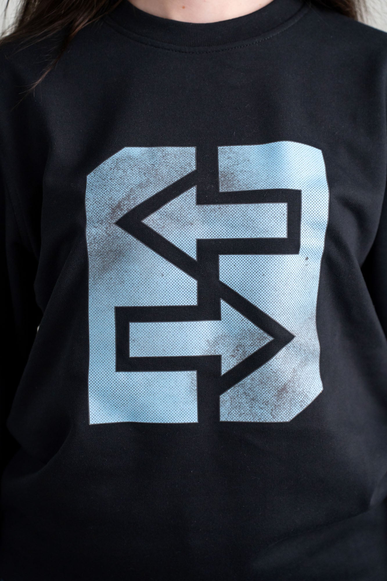 Eine Frau trägt ein schwarzes Sweatshirt mit einem blauen Logo, inspiriert von griechischer Mythologie und Geschichte.
