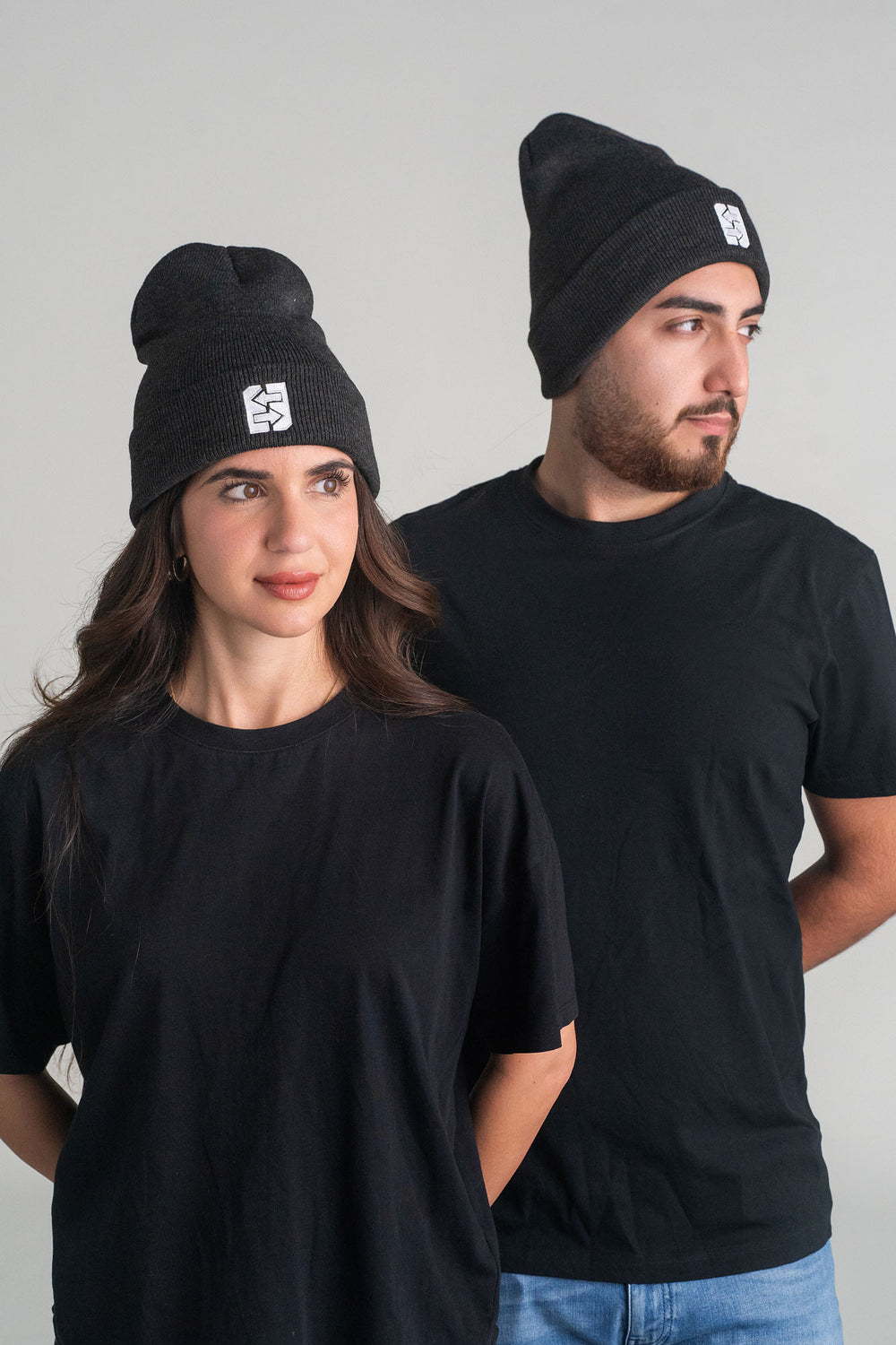 Ein Mann und eine Frau tragen schwarze T-Shirts und schwarze Mützen, lächelnd in einer urbanen Umgebung.
